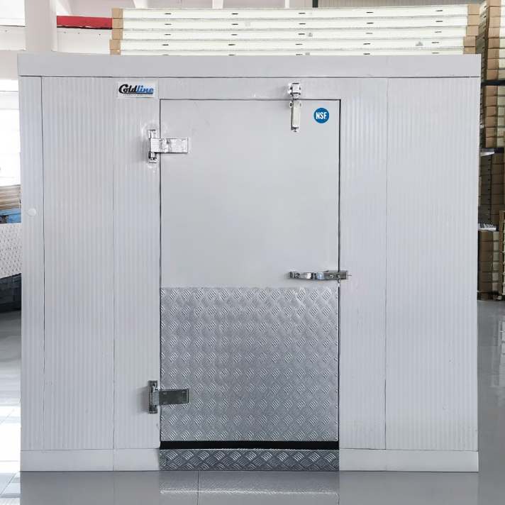 A wide walk-in refrigerator in the middle of a brightly lit warehouse. The front door of the walk-in is closed.