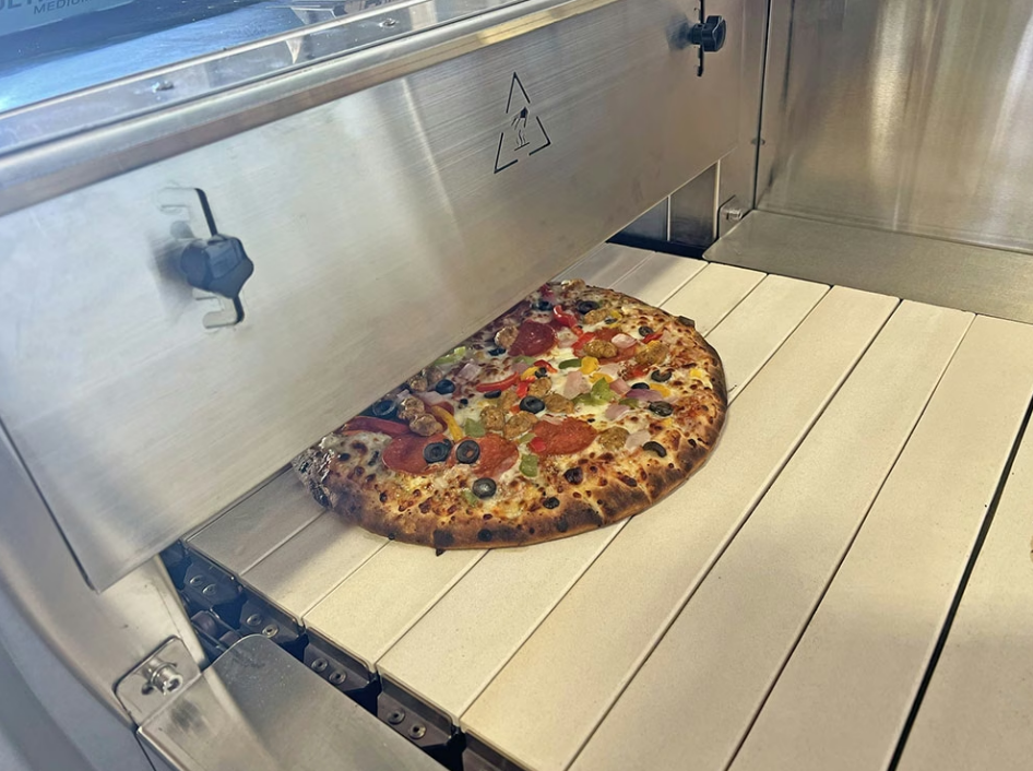 A cooked pizza with pepperoni and olives exiting a large metal commercial conveyor oven inside a restaurant.
