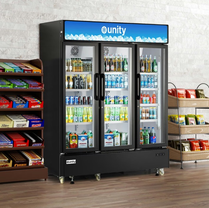 A commercial merchandise refrigerator with three glass doors full of drinks between food displays in a shop.